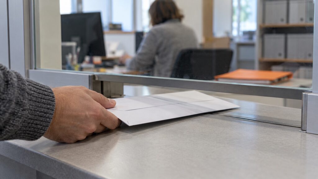 Ruka v sivom svetri podáva bielu zalepenú obálku cez okienko podateľne úradu. Za sklom je rozostrená kancelária s úradníčkou pri počítači a policami so šanónmi.